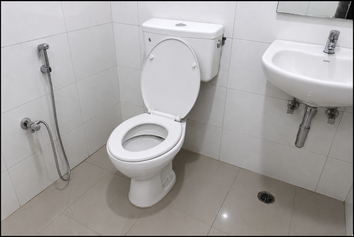 A white ceramic toilet with the lid open, shown in a tiled bathroom next to a white wall-mounted sink and a handheld shower attachment.